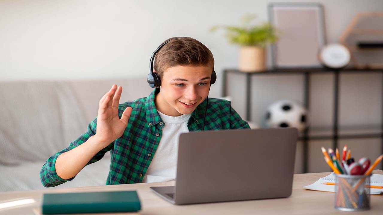 Positive teenager having video conference on laptop with teacher from home, wearing headset and waving at screen, free space